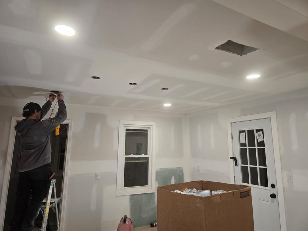 Person installing ceiling lights in a partially finished room with drywall and a cardboard box.