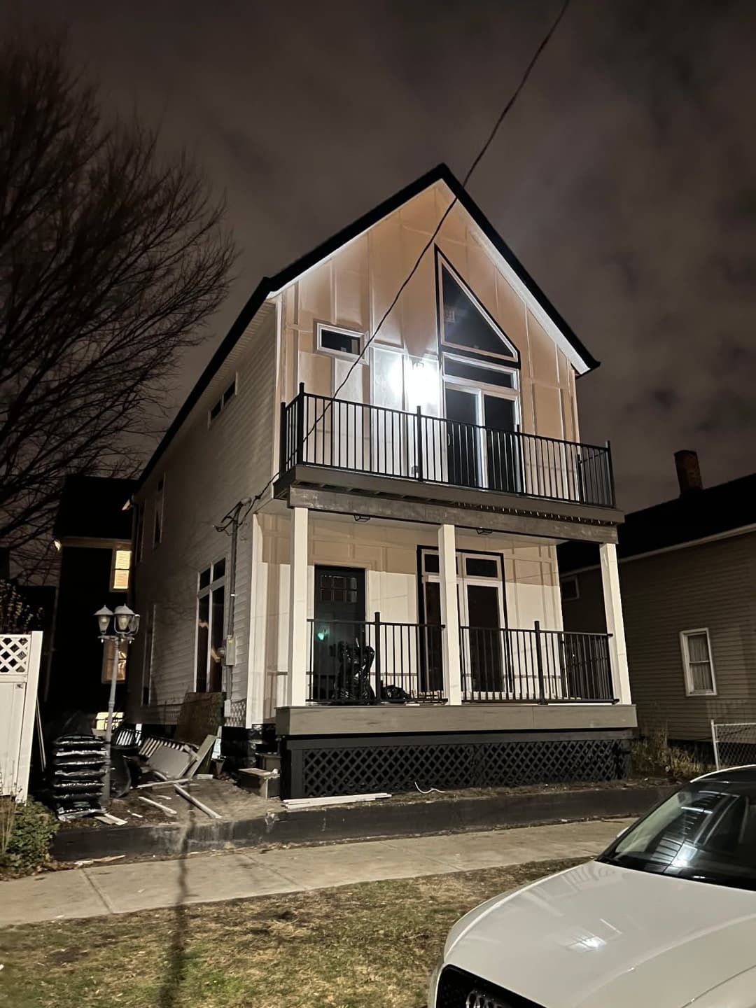 Two-story house at night with illuminated balcony and parked car in foreground.