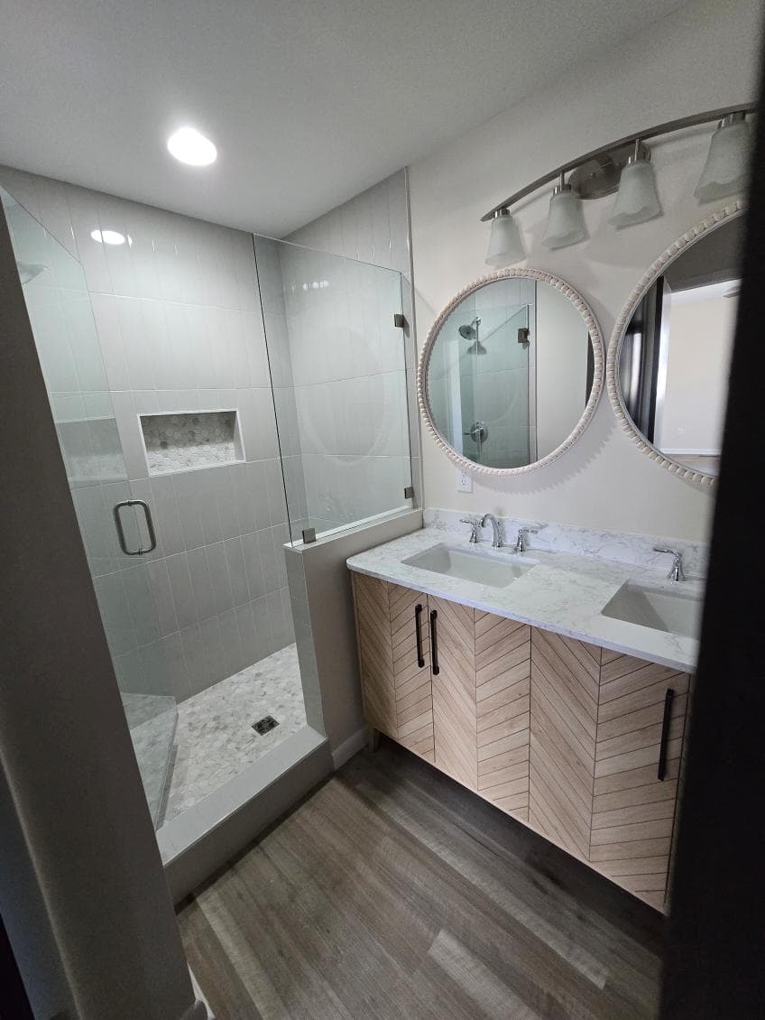 Modern bathroom with glass shower, double round mirrors, and light wood cabinetry.