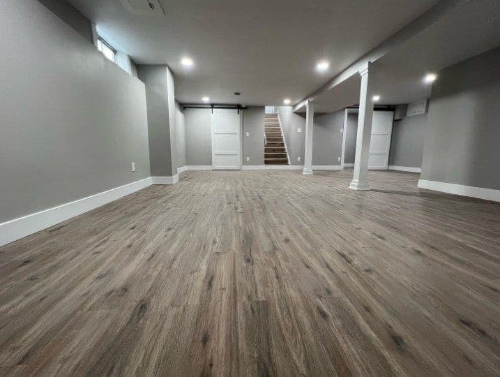 Spacious gray-finished basement with wooden flooring and white support pillars.